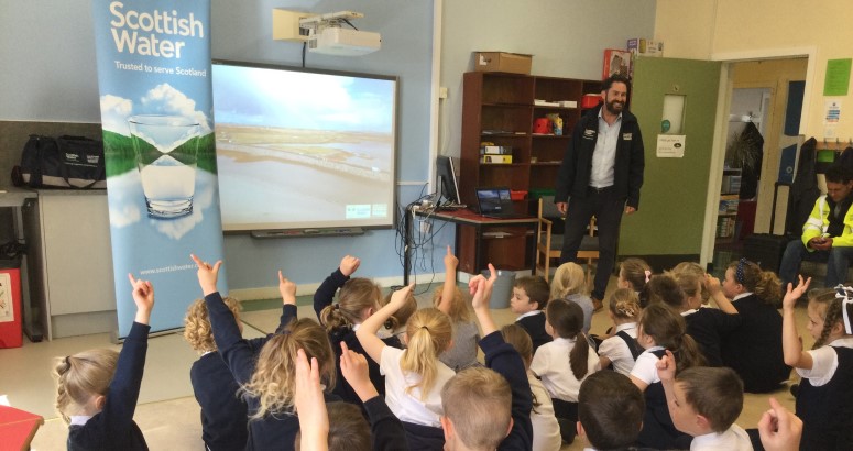 Benbecula School Visit A group of schoolchildren sitting on the floor, raising their hands towards an adult presenter near a screen displaying "Scottish Water" logo and landscape. The setting is an educational classroom.