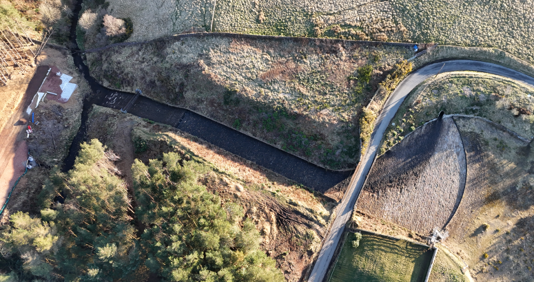 Gladhouse Reservoir Spillway Channel and Road Bridge Aerial view of a curved road passing through a rural landscape with patches of grass, trees, and a small building. A bridge crosses a narrow stream.