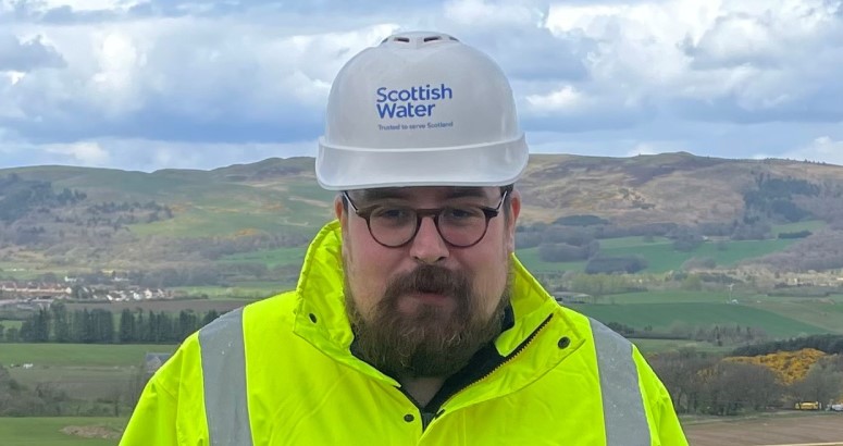 Neil Fox Graduate Quantity Surveyor Blog Person with beard posing with branded Scottish Water hard hat and hi-vis PPE, with lots of green hills in the background