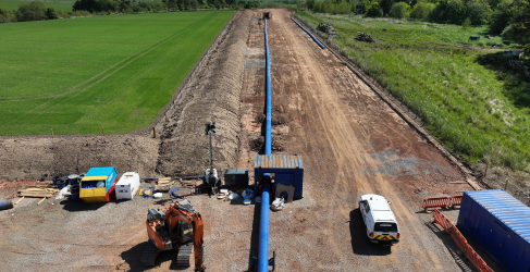 Newfield Project Progress Aerial view of a rural pipeline construction site with machinery, including an excavator and various vehicles, alongside materials laid out on cleared land under a clear sky.