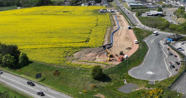 Newfield Project Progress Aerial view of a vibrant landscape showing a large bright yellow rapeseed field alongside roads with moving vehicles and construction equipment near a developing area. Rolling hills and greenery are visible in the background under a clear sky.