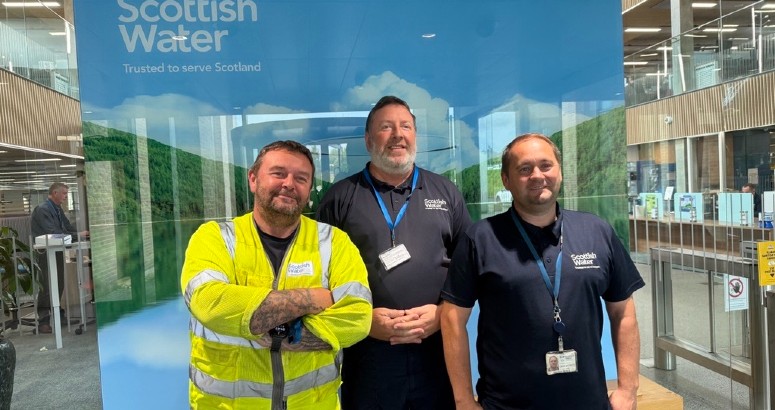 Field Response Clyde Team Celebrates Long-Service Three employees standing in front of a Scottish Water banner inside an office building. The person on the left is wearing a high-visibility vest, and the other two are in casual work attire, all smiling at the camera.